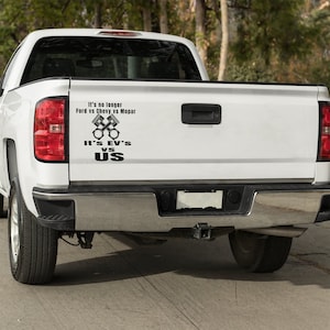 Puede incluir: Camioneta blanca con una calcomanía negra en la puerta trasera que dice "It's no longer Ford vs Chevy vs Mopar It's EV's vs US" con un gráfico de pistones cruzados.