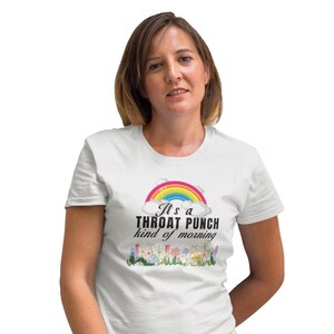 May include: A white short-sleeved t-shirt featuring a graphic of a colorful rainbow above clouds, the black text "It's a THROAT PUNCH kind of morning," and a row of various colorful flowers along the bottom hem of the printed design.
