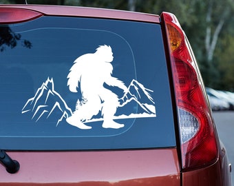 Könnte beinhalten: Weißes Vinyl-Aufkleber von Bigfoot, der vor einer Bergkette läuft. Das Design ist auf der Heckscheibe eines roten Fahrzeugs angebracht. Die Bigfoot-Silhouette ist detailliert, mit einem zotteligen Aussehen. Die Berge sind stilisiert mit scharfen Gipfeln.