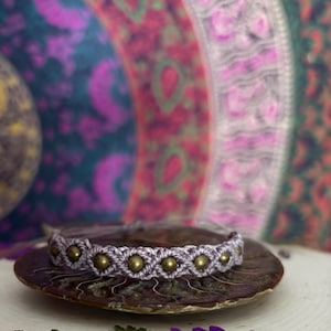 Può includere: Un braccialetto in macramè lavanda con perline dorate, appoggiato su una superficie con motivo marrone e bianco. Il braccialetto è realizzato con una tecnica di annodatura e presenta una serie di perline dorate distanziate uniformemente lungo la lunghezza del braccialetto.