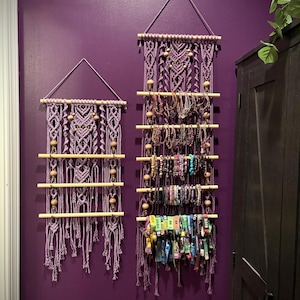 Könnte beinhalten: Zwei Makramee-Armband-Organizer in Lila hängen an einer lila Wand. Die Organizer haben Holzdübel und Perlen, wobei Armbänder am größeren Organizer ausgestellt sind. Das Makramee hat eine dreieckige Spitze.
