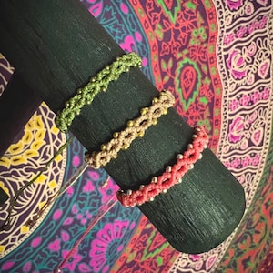 May include: Three friendship bracelets made with colourful string and beads. The bracelets are displayed on a wooden dowel rod. The bracelets are green, beige, and pink.