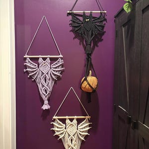 May include: Three macrame bat wall hangings in varying colors: lavender, black, and cream. Each bat is suspended from a wooden dowel and a triangular macrame hanger. A small pumpkin hangs from the black bat. The bats are displayed on a purple wall.