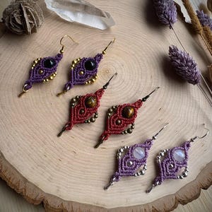 May include: Three pairs of macrame earrings featuring diamond-shaped woven cord patterns. The top pair is purple with black beads and gold-tone accents, the middle is red with tiger eye stones and bronze beads, and the bottom is light purple with moonstone-like gems and silver-tone beads.