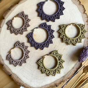 May include: Five circular hoop earrings featuring intricate macrame knotting in shades of purple, tan, and olive green. Each earring has a metallic hoop frame with a starburst-like knotted pattern extending outward from the center of the hoop.