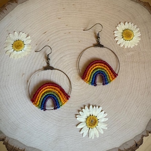 Könnte beinhalten: Handgefertigte Regenbogen-Ohrringe mit Makramee-Design. Die Ohrringe zeigen einen farbenfrohen Regenbogenbogen, der an einem silbernen Reifen befestigt ist. Die Ohrringe werden auf einer Holzoberfläche mit drei weißen Gänseblümchen präsentiert.
