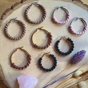 Könnte beinhalten: Eine Auswahl handgefertigter Creolen in verschiedenen Farben, darunter Braun, Rosa und Schwarz, auf einer Holzoberfläche präsentiert. Die Ohrringe weisen ein gewebtes oder geknotetes Design mit gold- und silberfarbenen Metallakzenten auf. Trockenblumen und ein rosa Kristall sind ebenfalls vorhanden.