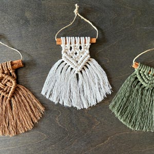 Puede incluir: Tres tapices de macramé en marrón, blanco y verde. Cada pieza presenta una clavija de madera y un diseño anudado con flecos. La pieza marrón tiene un nudo de diamante, la blanca tiene un diamante central y la verde tiene un diseño de semicírculo.