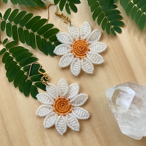 May include: A pair of white daisy earrings with gold hooks. The daisies have a yellow center and are made of macrame.