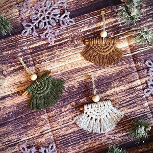 May include: Handmade macrame Christmas decorations in brown, green, and white, each with a wooden bead and cinnamon stick detail. Glittering purple snowflake ornaments and evergreen branches add to the festive display.