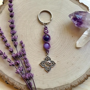 May include: A purple macrame keychain with amethyst and a silver charm with an Om symbol. The keychain is displayed on a wooden surface with lavender sprigs and a crystal. The color palette includes shades of purple, silver, and natural wood tones.