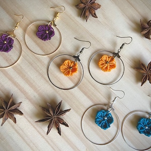 May include: A set of six hoop earrings with crocheted flower charms. The earrings are available in three colours: purple, orange, and blue. The hoops are made of gold, silver, and bronze metal.