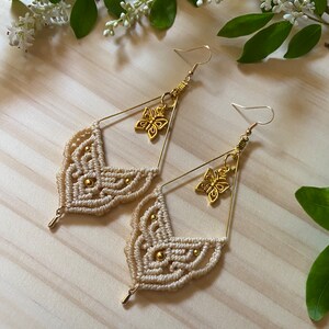 Pode incluir: Um par de brincos de macramé feitos à mão com um amuleto de borboleta dourado. Os brincos são feitos com cordão de cor creme e têm um design geométrico.