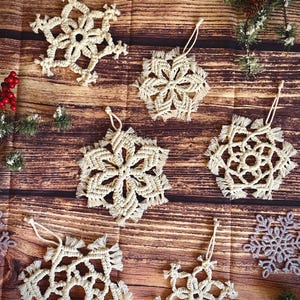 May include: A collection of handmade, off-white macrame snowflake ornaments with hanging loops. The snowflakes are arranged on a wooden surface, with decorative elements like red berries and pine branches. These ornaments are perfect for holiday decor.
