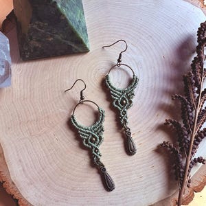 May include: Handmade macrame earrings in a soft green hue, featuring a circular design with intricate knotting and a teardrop-shaped pendant. The earrings are displayed on a wooden surface, showcasing a bohemian style.