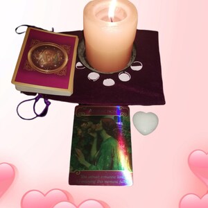 May include: A purple velvet altar cloth with a white candle in a metal holder, a white heart-shaped crystal, and two oracle cards. One card is gold and purple with a picture of a woman and the other card is iridescent with a picture of a couple in a garden. The text on the iridescent card reads "You attract romantic love by enjoying this moment fully."