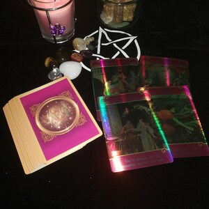 May include: A pink candle in a glass jar with a metal cage, surrounded by crystals and tarot cards. The cards are iridescent and have text that reads "You've already met the romantic partner you seek."