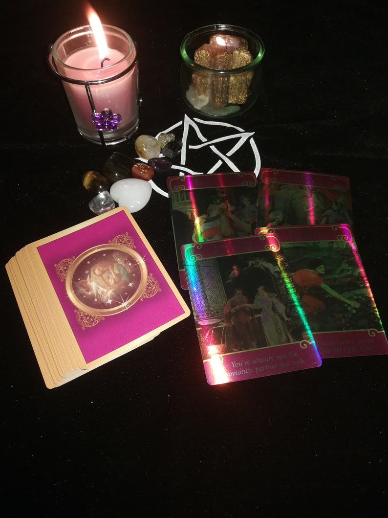 May include: A pink candle in a glass jar with a metal cage, surrounded by crystals and tarot cards. The cards are iridescent and have text that reads "You've already met the romantic partner you seek."