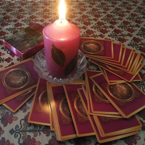 May include: A lit pink candle with a leaf design sits on a glass plate surrounded by a spread of tarot cards with gold trim and a burgundy background.