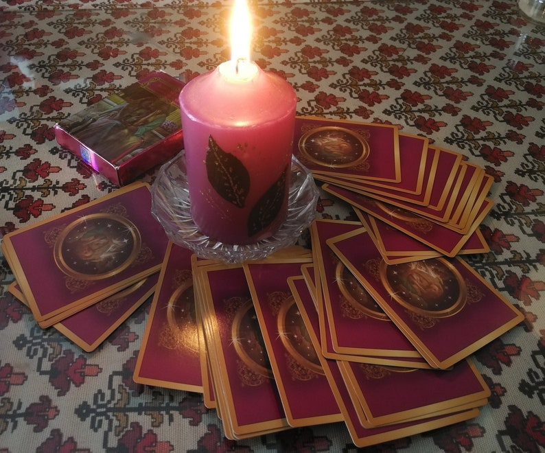 May include: A lit pink candle with a leaf design sits on a glass plate surrounded by a spread of tarot cards with gold trim and a burgundy background.