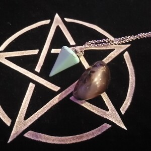 May include: A silver chain necklace with a light blue crystal pendulum and a dark gray stone on a black background with a white pentagram.