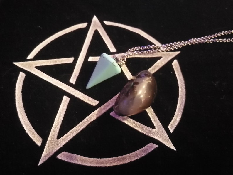 May include: A silver chain necklace with a light blue crystal pendulum and a dark gray stone on a black background with a white pentagram.