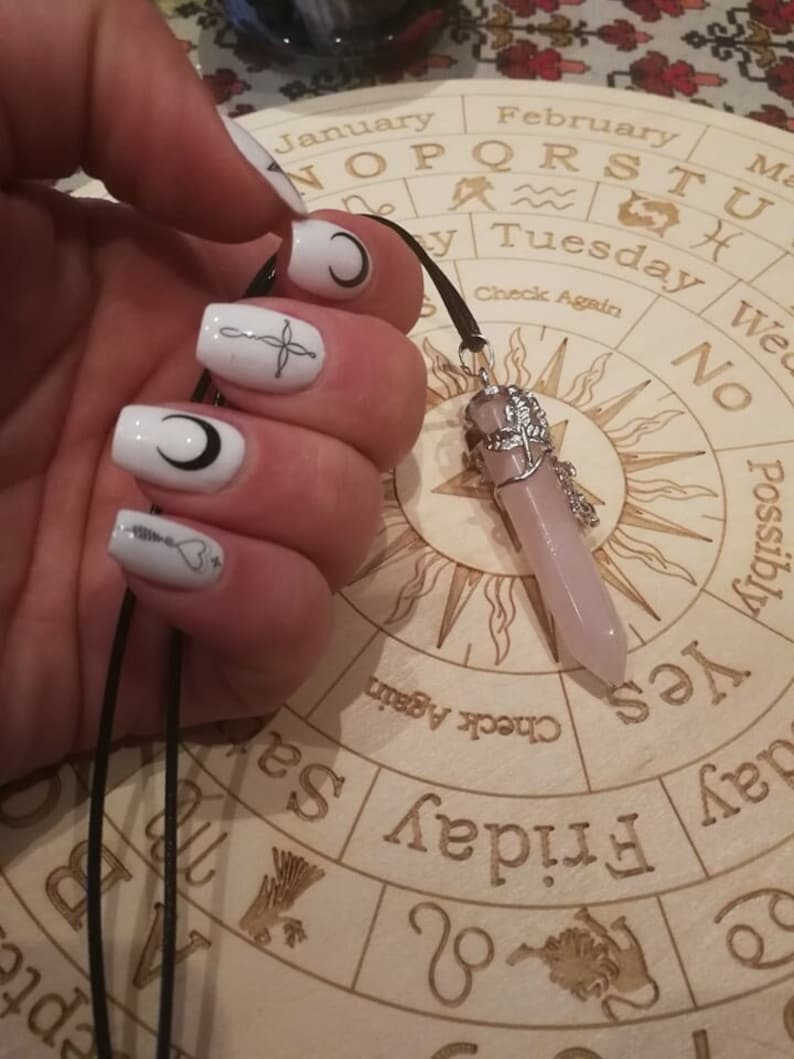 May include: A wooden Ouija board with a black cord and a pink crystal pendant. The board has a circular design with the letters of the alphabet, numbers, and the words "yes", "no", "goodbye", and "check again".