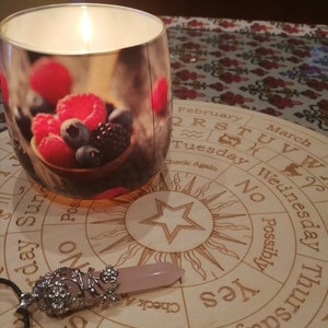 May include: A wooden Ouija board with a star in the center and a pink crystal pendulum. The board has the letters of the alphabet, numbers, and the words "yes", "no", "possibly", and "check again".