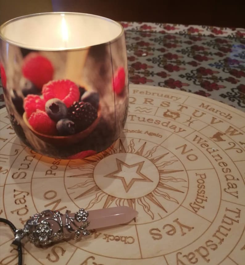 May include: A wooden Ouija board with a star in the center and a pink crystal pendulum. The board has the letters of the alphabet, numbers, and the words "yes", "no", "possibly", and "check again".