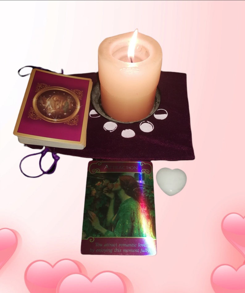 May include: A purple velvet altar cloth with a white candle in a metal holder, a white heart-shaped crystal, and two oracle cards. One card is gold and purple with a picture of a woman and the other card is iridescent with a picture of a couple in a garden. The text on the iridescent card reads "You attract romantic love by enjoying this moment fully."