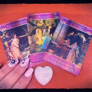 May include: Three oracle cards with colourful illustrations and text. The cards are laid out on a wooden surface. The text on the cards reads "You Deserve Love", "Retreat", and "This Could Be The One".