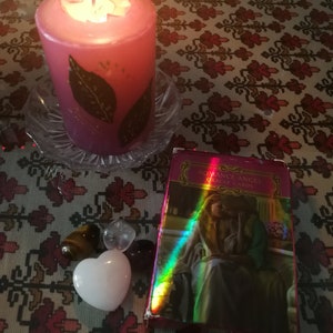 May include: A pink candle with gold leaf accents is burning on a glass plate.  There are four crystals and a white heart-shaped stone on a patterned tablecloth.  A deck of Romance Angel Oracle Cards is on the table.