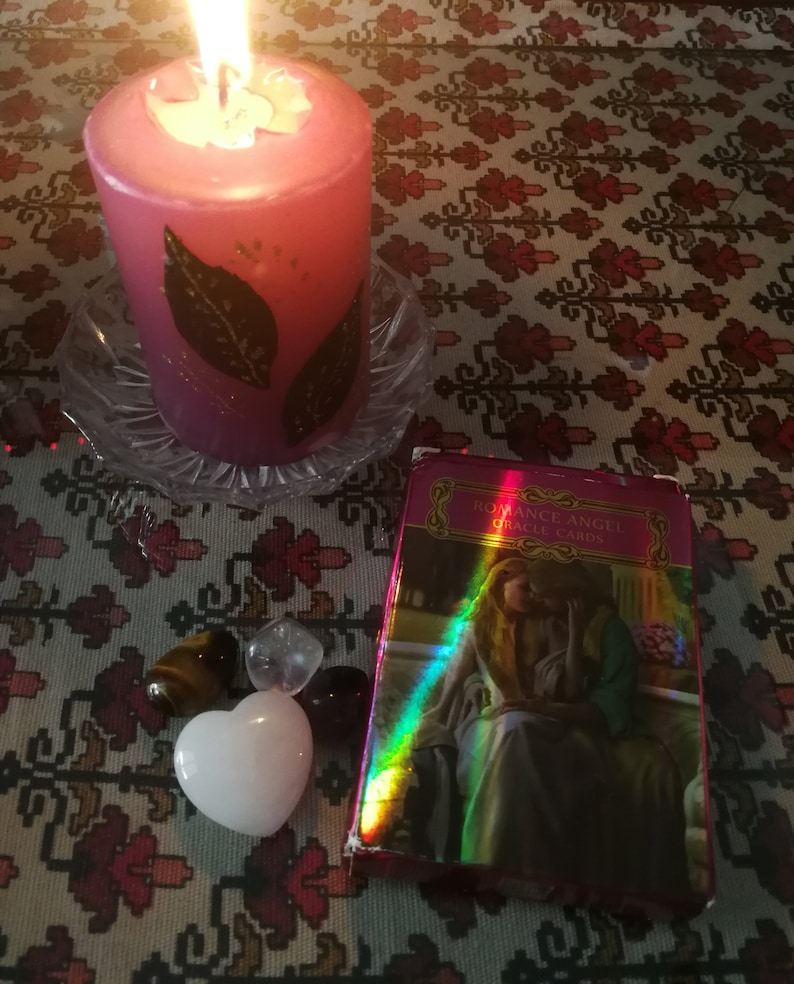 May include: A pink candle with gold leaf accents is burning on a glass plate.  There are four crystals and a white heart-shaped stone on a patterned tablecloth.  A deck of Romance Angel Oracle Cards is on the table.