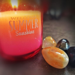 May include: A red glass candle jar with a yellow and orange candle inside. The candle has the text "Welcome Summer Sunshine" on the side. There are four smooth stones next to the candle, two orange, one black, and one dark brown.