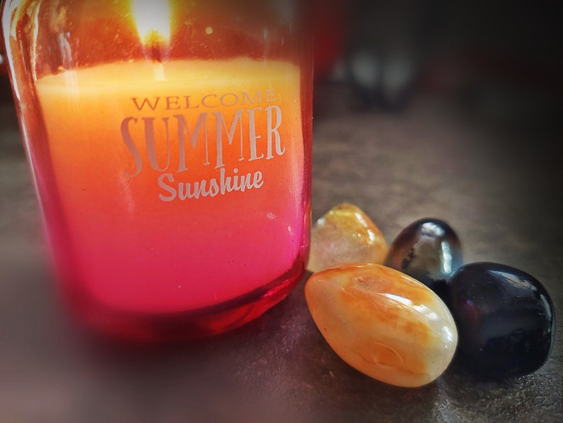 May include: A red glass candle jar with a yellow and orange candle inside. The candle has the text "Welcome Summer Sunshine" on the side. There are four smooth stones next to the candle, two orange, one black, and one dark brown.