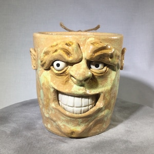 May include: A ceramic mug shaped like a face with a wide, toothy grin. The mug is glazed in a light brown color with green and yellow accents. The eyes are white with black pupils.
