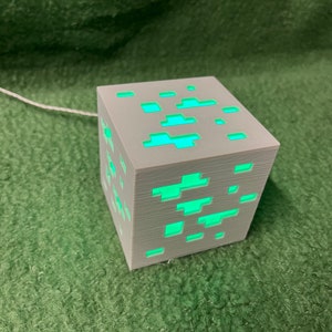 Könnte beinhalten: Eine graue, würfelförmige Lampe mit leuchtend grünen Akzenten, die an einen Videospielblock erinnert. Die Lampe hat eine Reihe rechteckiger und quadratischer Aussparungen, die ein hellgrünes Licht aussenden. Ein weißes Kabel erstreckt sich von der Lampe.