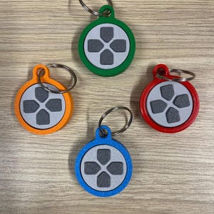 Könnte beinhalten: Vier kreisförmige Schlüsselanhänger mit einem zentralen grauen Steuerkreuz-Design. Jeder Anhänger hat eine eigene Außenringfarbe: Grün, Orange, Blau und Rot. Jedes Stück enthält einen silbernen Metallring an der oberen Öse.