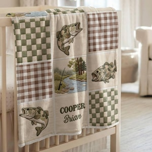 May include: A baby blanket with a fishing theme, draped over a crib. The blanket features squares of green and brown checkered patterns, embroidered fish, and a river scene. The name "Cooper Brian" is embroidered on the blanket.