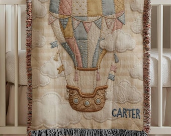 May include: A beige baby blanket with a colorful hot air balloon design. The balloon features pastel pink, blue, and yellow panels, with a brown basket and decorative flags. The name "CARTER" is embroidered in blue. The blanket is fringed.