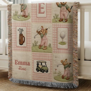 May include: A woven throw blanket featuring a grid pattern with illustrations of teddy bears playing golf, a golf bag, a golf cart, and a golf ball on a tee. The blanket includes the name Emma Rose in pink text and a large letter E.
