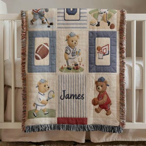 May include: A beige and blue sports-themed baby blanket with a fringe. The blanket features squares with sports illustrations, including hockey, baseball, football, basketball, and tennis. Teddy bears in sports uniforms are also featured. The name "James" is embroidered on the blanket.