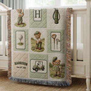 May include: A beige and green golf-themed baby blanket with a patchwork design. The blanket features illustrations of teddy bears playing golf, golf clubs, and a golf cart. The name "Harrison" and the date "June 26, 2026" are printed on the blanket.