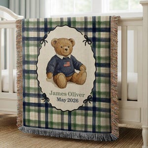Peut inclure: Une couverture pour bébé tissée avec un motif à carreaux vert, bleu et crème. La couverture présente un motif ovale central avec un ours en peluche portant un pull avec un drapeau américain. Le texte "James Oliver May 2026" est en dessous.