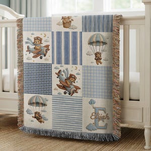 Peut inclure: Un plaid tissé présentant un motif patchwork avec des rayures bleues et blanches, des motifs vichy et des illustrations d'ours en peluche en tenue d'aviateur pilotant des avions et faisant du parachute. Le plaid inclut le nom Elijah et un grand E.