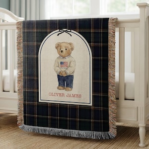 May include: A navy blue and green plaid throw blanket with a cream-colored rectangular design featuring a teddy bear wearing a sweater with an American flag. The blanket has a fringed edge and the name "OLIVER JAMES" is printed at the bottom.