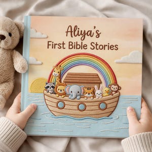 May include: A children's book titled "Aliya's First Bible Stories" with an illustration of Noah's Ark, a rainbow, and various animals. The book is held by a person, and a teddy bear is next to it. The cover has a light blue border and a soft, pastel colour scheme.