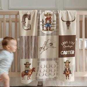 May include: A beige and brown patchwork baby blanket with cowboy-themed illustrations. The blanket features images of a cowboy bear, a bull skull, horseshoes, and the text "Long Live Cowboys CARTER". The blanket is draped over a wooden crib.