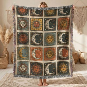 May include: Woven throw blanket featuring a repeating pattern of suns and moons in square frames. The celestial design is in shades of orange, yellow, blue, and cream, with a fringed edge. The blanket is held up by a person.