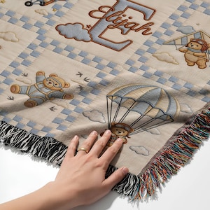 May include: A woven throw blanket featuring a beige background with a blue and white checkered pattern. The design includes illustrations of teddy bears wearing aviator goggles, flying with parachutes, kites, and planes, and the name Elijah.